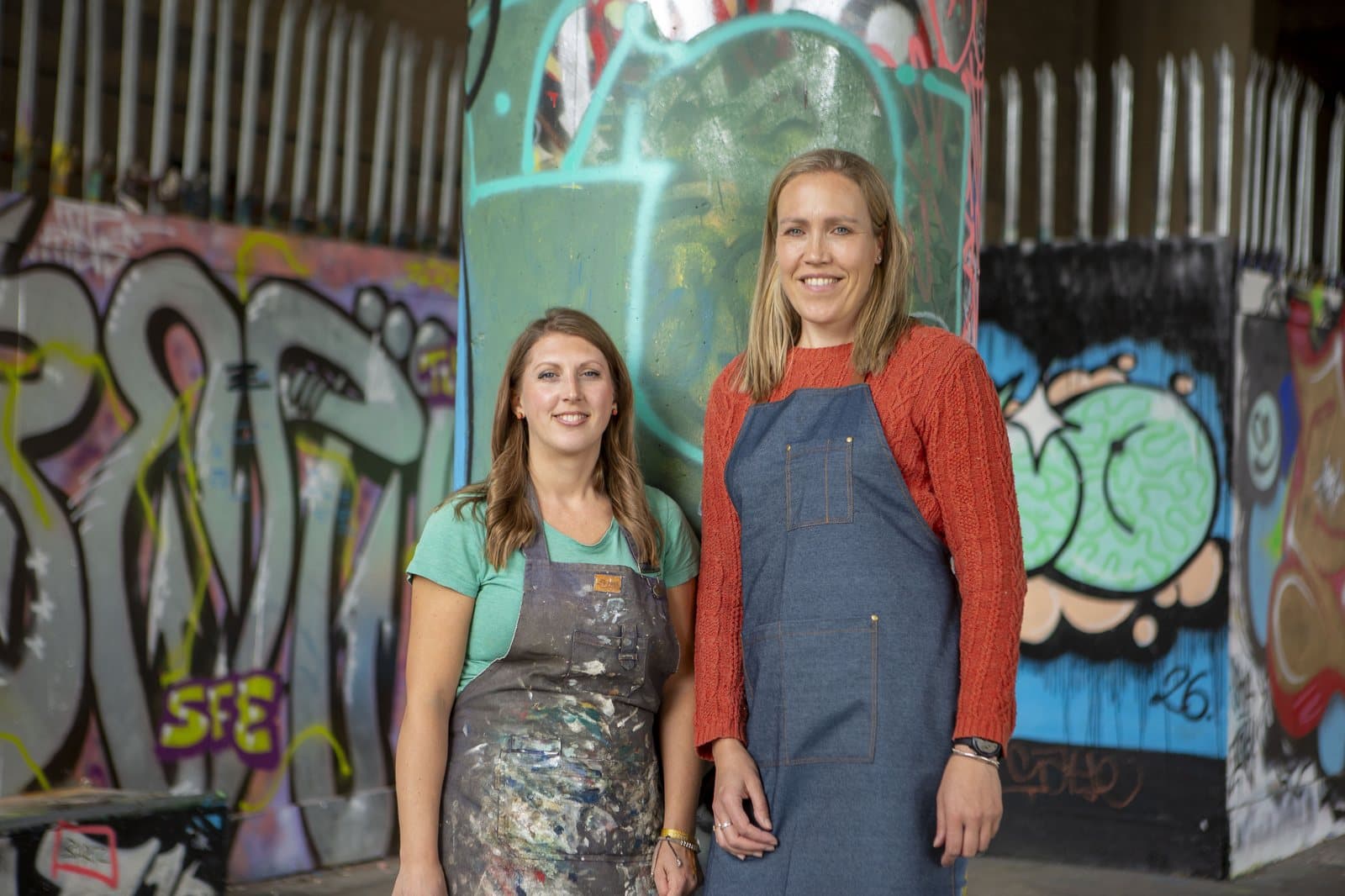 Ally Booker and Gemma Broad, co-founders of We Are Repairs, standing in work aprons in front of a graffiti wall